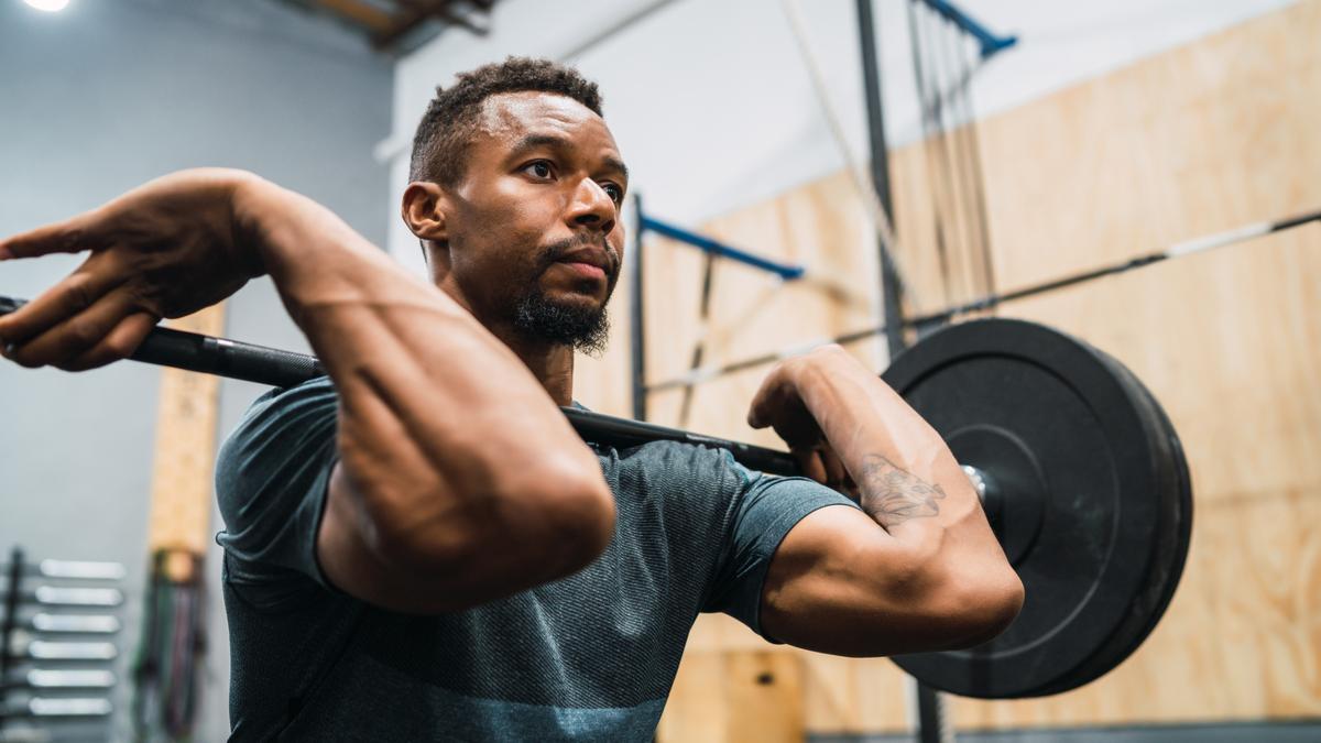 A person wears a blue-grey t-shirt and holds a loaded barbell in a front rack position.