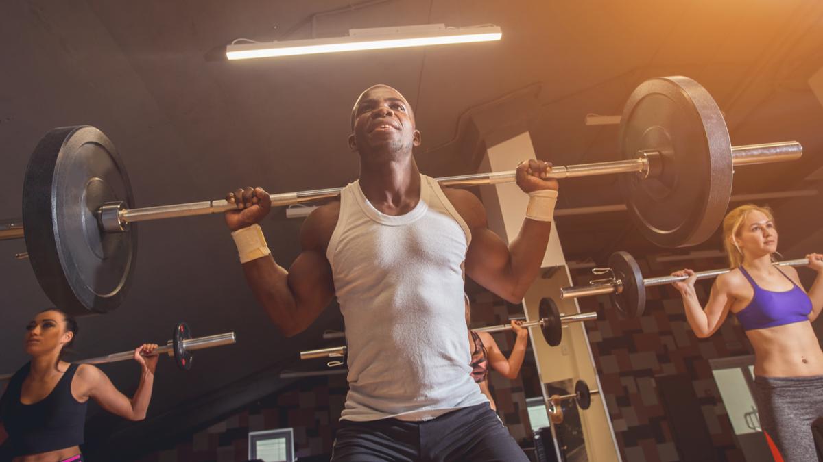 A person wearing a white tank top prepares to back squat with three people behind them, also preparing to back squat.